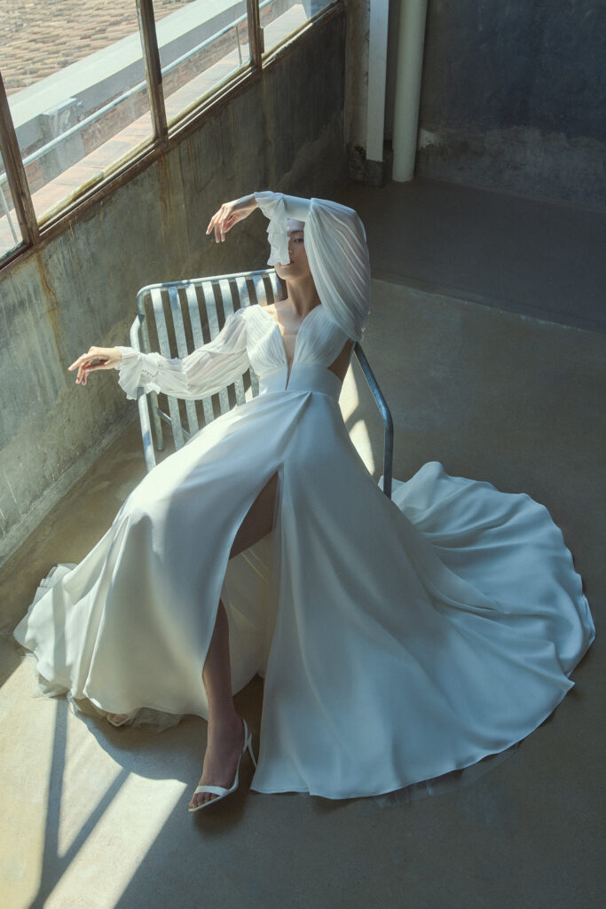 femme assise habillée d'une robe de mariée fluide raffinée à fente et à manches longue en mousseline plissé